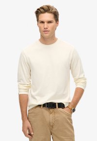 Lange mouw crèmekleurig shirt van zachte stof met een ronde halslijn, gecombineerd met tan broek en een zwarte riem, met een gouden horloge om de pols.