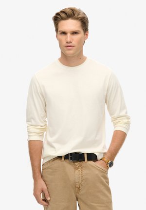 Camisa de manga larga color crema en tejido suave con un escote redondeado, combinada con pantalones color caqui y un cinturón negro, luciendo un reloj dorado en la muñeca.