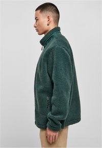 Giacca in pile verde scuro con colletto alto, tasca laterale con zip e superficie texturizzata. Presenta una vestibilità comoda e maniche lunghe.