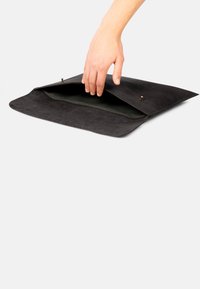 Schwarze Wildledertasche im Umschlagstil mit glatter Textur, ausgestattet mit einem grünen Innenfutter und einem Druckknopfverschluss. Eine Hand greift in die offene Tasche.