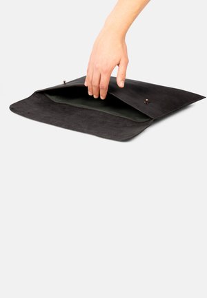 Schwarze Wildledertasche im Umschlagstil mit glatter Textur, ausgestattet mit einem grünen Innenfutter und einem Druckknopfverschluss. Eine Hand greift in die offene Tasche.