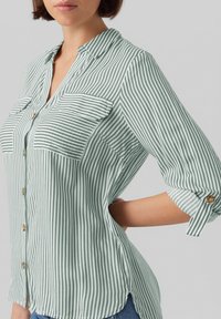 Green and white striped blouse with a buttoned front, two chest pockets, and rolled sleeves. Soft fabric with a smooth texture.