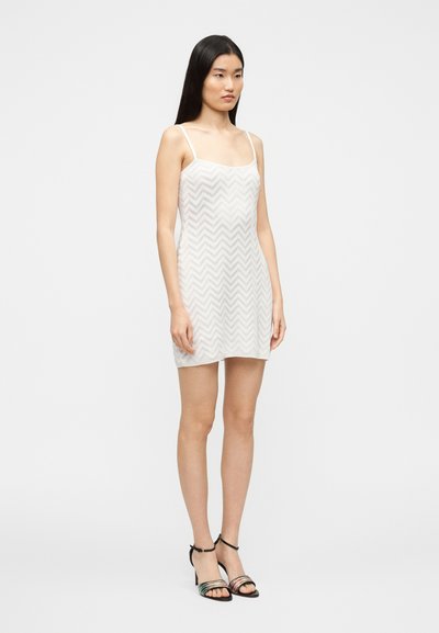 Robe mini blanche sans manches à motif chevron, dotée de fines bretelles et d'une texture douce, associée à des sandales à talons noires.