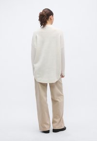 Šviesiai smėlio spalvos didelio dydžio megztinis su tekstūruotu paviršiumi, aukštu kaklu ir nuleistais pečiais, derinamas su smėlio spalvos plačiomis kelnėmis.