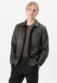 Dunkelgrüne Lederjacke mit Knopfverschluss, Brusttasche und glatter Textur. Getragen über einem grauen Hemd und schwarzen Cordhosen.