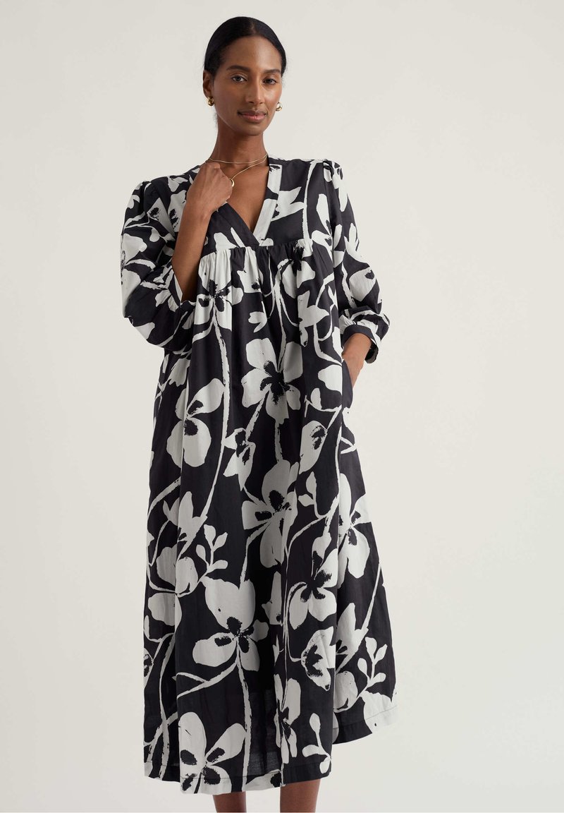 Femeie purtând o rochie midi neagră cu imprimeu floral alb supradimensionat, mâneci lungi bufante, decolteu în V și buzunare frontale, pozând pe un fundal simplu.