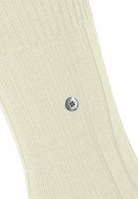 Jasno kremowe skarpetki w prążki o gładkiej strukturze, z okrągłą srebrną metką logo na środku. Materiał wydaje się miękki i elastyczny.