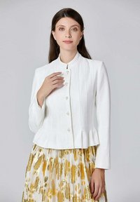 Veste légère blanche avec un col haut, fermeture à boutons, tissu à texture subtile et ourlet en peplum. Portée avec une jupe à motif doré.