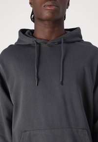 Dunkelgrauer Hoodie aus strukturiertem Stoff, mit Kordelzug an der Kapuze und einer Fronttasche. Einfaches Design ohne sichtbare Muster oder Akzente.