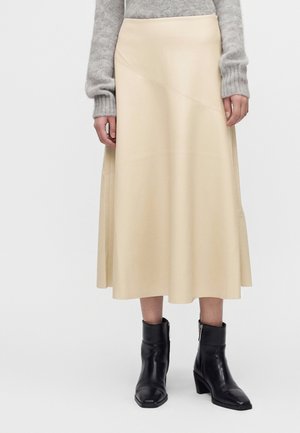 Femme portant une jupe midi beige, un pull gris à manches longues et des bottines noires à talons, debout devant un fond blanc uni.