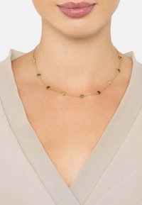 Guldtonet halskæde med cirkulære led og små farvede accenter. Let vægt kæde, der hviler tæt på halsen mod en beige top.