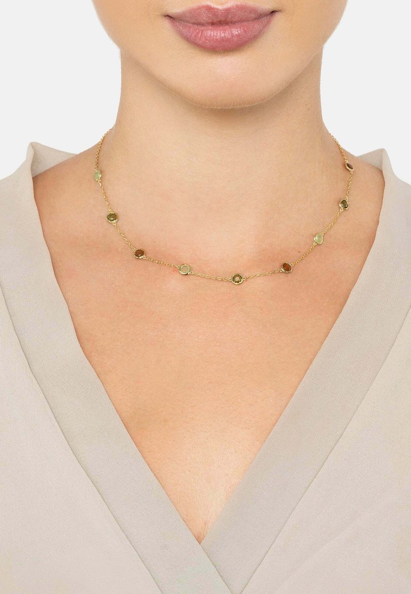 Guldtonet halskæde med cirkulære led og små farvede accenter. Let vægt kæde, der hviler tæt på halsen mod en beige top.