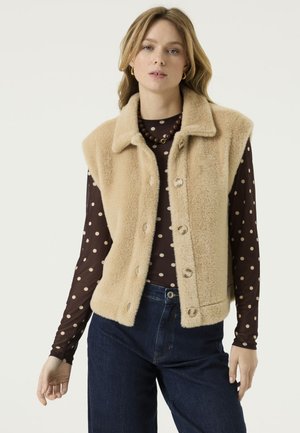 Mujer con chaleco sin mangas de forro polar beige abotonado sobre una camisa marrón de manga larga con lunares y vaqueros azul oscuro sobre un fondo liso.