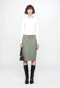 Haut blanc à manches longues associé à une jupe olive de longueur genoux avec des poches. Bottes noires montant aux genoux, tenant un petit sac à main marron.