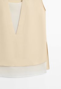 Beige sleeveless V-neck top layered over a white underlayer with side slits on a plain white background.