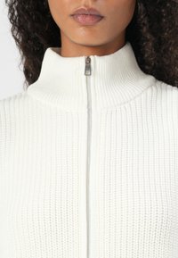 Pull en maille blanche texturée avec un col montant et une fermeture éclair argentée. Le tissu présente un motif côtelé et un ajustement près du cou.