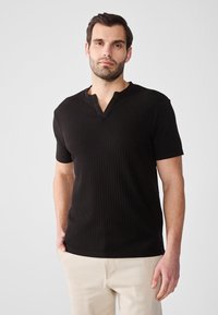 Sort ribbet skjorte med kortærmer og placket halsudskæring. Stoffet ser tekstureret ud med let stræk og er sat sammen med lyse bukser.