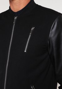 Chaqueta bomber negra con mangas de cuero de contraste, que incluye múltiples bolsillos con cremallera y un diseño clásico de cuello.