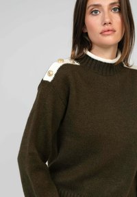 Pull en maille vert foncé avec un col montant. Présente des accents blancs aux épaules et des boutons dorés pour les détails. Texture douce et coupe décontractée.