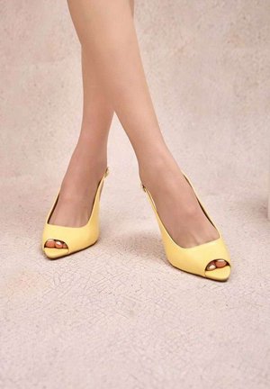 Jambes croisées aux chevilles portant des escarpins slingback à bout ouvert jaunes sur un sol texturé beige clair.