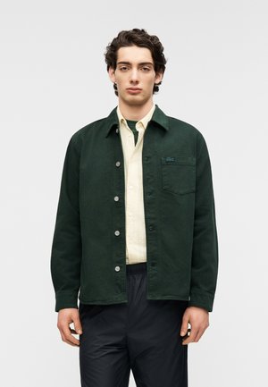 Giacca verde scuro in tela con chiusura a bottoni, colletto e taschino sul petto. Indossata sopra una camicia giallo chiaro, abbinata a pantaloni neri.