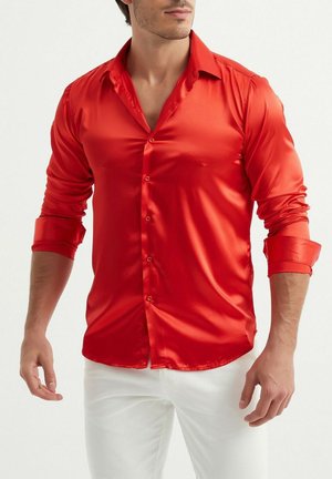 Chemise longue à manches longues en satin rouge avec un devant à boutons, un col pointu et des poignets retroussés. Texture lisse, design ajusté, portée avec un pantalon blanc.