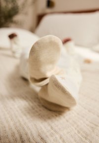 Kreemikujulised sokid, millel on peened tähemustrit, lebamas tekstuurse heledabežika tekil. Pehme kangas, ümardatud varba kujundus ja nähtav õmblus.