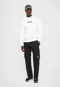 Felpa bianca con etichetta nera, pantaloni cargo neri con tasche laterali e sneakers nere. Il cappellino è grigio scuro con un visiera decorata.