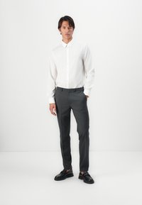 Hvit skjorte med lange ermer, grå slim-fit bukser og svarte loafers, som står foran en helt hvit bakgrunn.