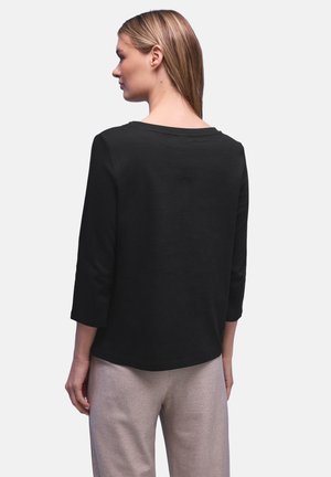 Mujer con cabello rubio lacio, lleva una blusa negra de manga 3/4 y pantalones texturizados de color claro, de espaldas sobre un fondo liso.