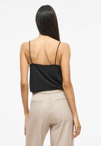 Kobieta z długimi włosami, ubrana w czarny bezrękawnik i beżowe spodnie z wysokim stanem, stojąca tyłem do kamery.