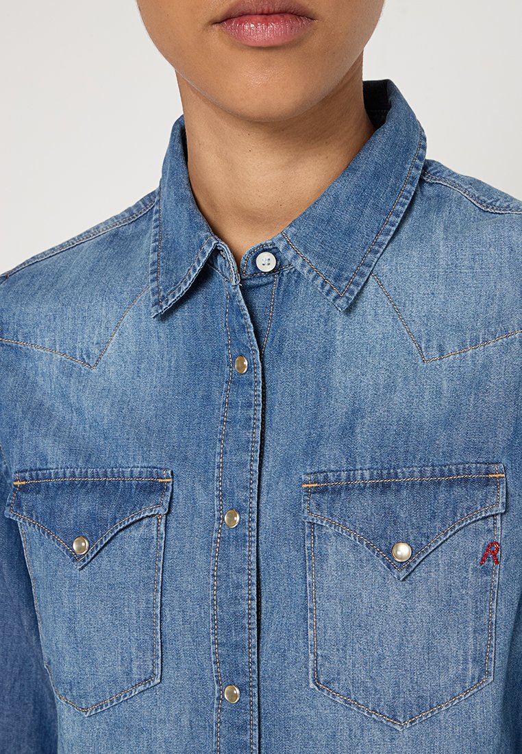 Camisa de denim con cuello clásico, color azul, dos bolsillos en el pecho con botones a presión y costuras en contraste. Presenta bordado del logo en el bolsillo.