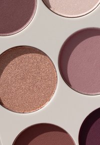 Round eyeshadow pans on a white palette feature shades of mauve, plum, and bronze, with textures ranging from matte to shimmer.