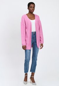 Roze gebreide cardigan met een V-hals en open voorkant, gecombineerd met lichtblauwe jeans en zilveren hoge hakken. Gestructureerde stof met subtiele glans.