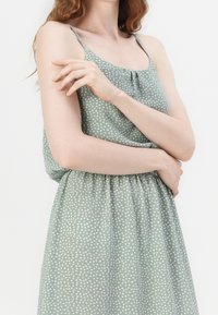 Vihreä kuviollinen mekko, jossa on ohuet hihnat, teksturoitu kangas ja kerätty vyötärö. Sisältää valkoisia pilkkuja koko matkalla.