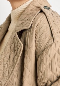 Chaqueta acolchada beige con cuello estructurado y detalles de botones, que presenta un patrón texturizado y un diseño de capas de tela.