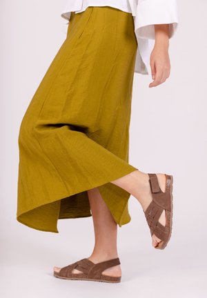 Mujer con sandalias marrones de tiras levantando un pie, vestida con una falda midi verde oliva y una camiseta blanca de manga larga.