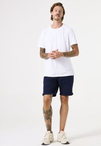 Weißes Baumwoll-T-Shirt, marineblaue Shorts mit umgeschlagenen Säumen und cremefarbene Sneaker. Zeigt Tätowierungen an den Armen und Beinen, steht vor einem neutralen Hintergrund.