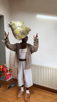 Groene en gele gestreepte dekbed in plastic folie, omhoog gehouden boven een persoon. Deze draagt een geruite shirt, een witte tanktop en grijze shorts.