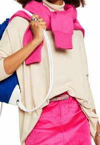 Beżowy sweter z różowym bluzą narzuconą na ramionach, w zestawieniu z różową spódnicą z korduroju i srebrnym paskiem. Widać niebiesko-białe plecak.