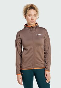 Bruine jas met rits en capuchon, oranje accenten, gestructureerde stof en een logo op de borst. Heeft een aansluitend ontwerp en zijzakken.