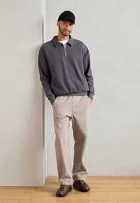 Harmaa neljänneksen vetoketjupullover, ribattua pintaa; vaaleanbeige rento-housut, näkyvät taskut; ruskeat nauhakenkä.