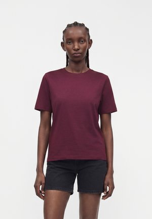 Femme aux cheveux tressés portant un T-shirt bordeaux uni à manches courtes et un short en jean noir, debout devant un fond blanc.