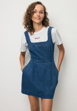 Młoda kobieta z kręconymi włosami uśmiecha się, nosząc białą koszulkę pod bezrękawną niebieską dżinsową sukienkę z kieszeniami, stojąc na jednolitym tle.