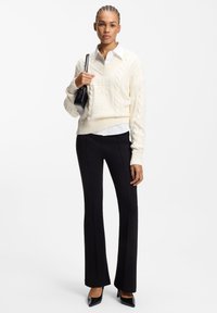 Cream cable-knit sweater with ribbed hem, layered over a white collared shirt. Black fitted trousers and pointed black heels complete the outfit.