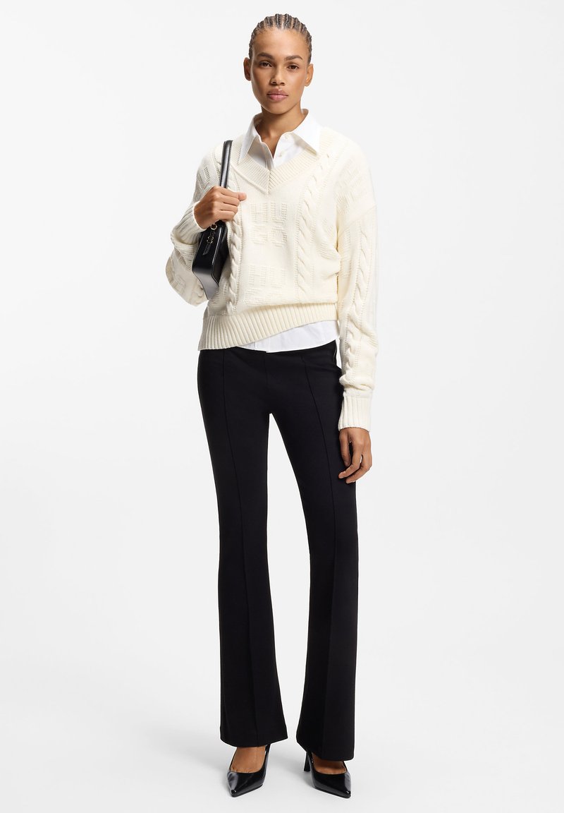 Cream cable-knit sweater with ribbed hem, layered over a white collared shirt. Black fitted trousers and pointed black heels complete the outfit.