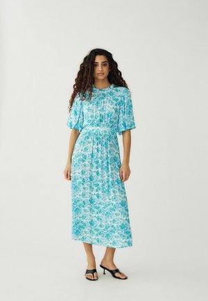 Femeie cu păr lung și creț purtând o rochie midi florală albastru-verde și sandale negre cu toc și degetele descoperite, stând în picioare pe un fundal simplu.