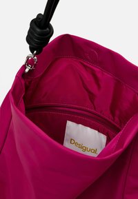 Innenseite einer pinken Nylon-Tasche mit einem Reißverschlussfach, glatter Textur und einem sichtbaren Etikett mit der Aufschrift "Desigual" in goldener Schrift.