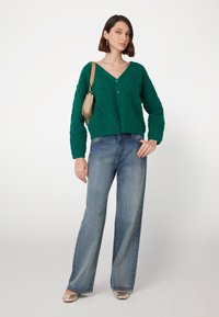 Cardigan verde a maglia con scollatura a V e bottoni, abbinato a jeans in denim blu a gamba larga e ballerine metallizzate, con una borsa semplice.