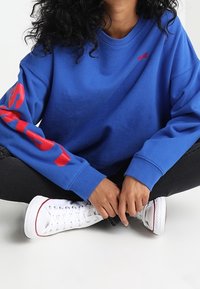 Niebieska bluza z czerwoną literą na rękawach, okrągły dekolt i luźny krój. Połączona z białymi wysokimi trampkami i czarnymi dżinsami.